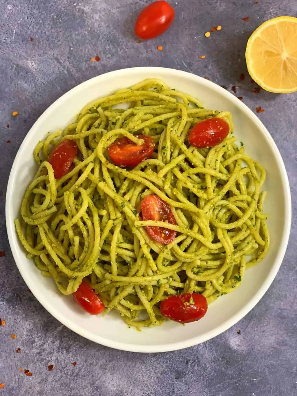 basil pesto spaghetti served in a white plate with tomaotes on top and lemon wedge on side