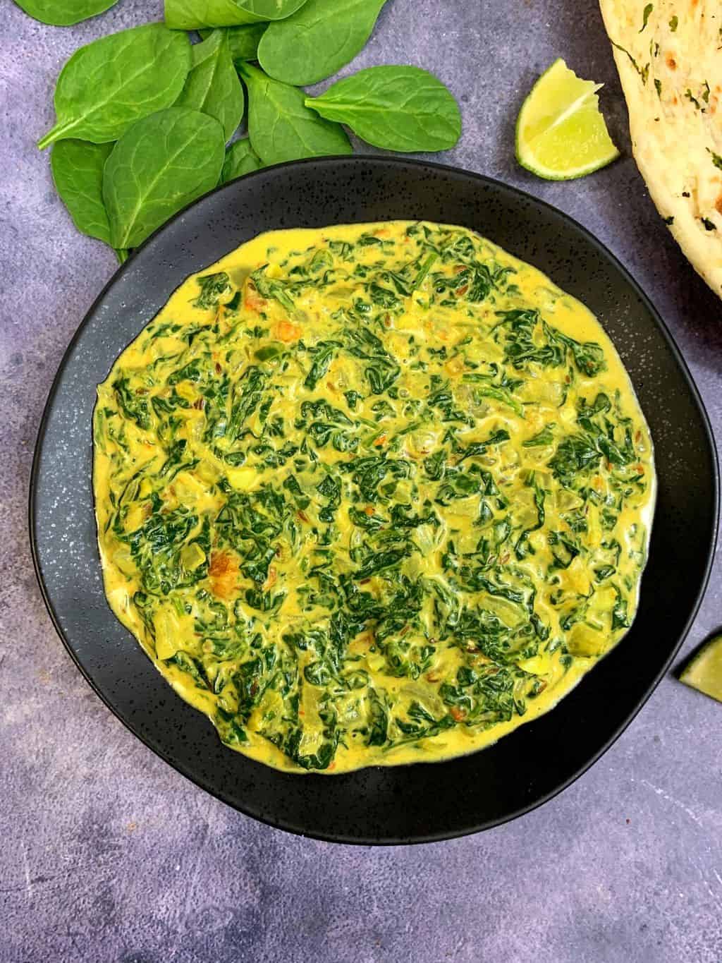 indian style creamed spinach served in a black plate with lemon wedges and naan on side