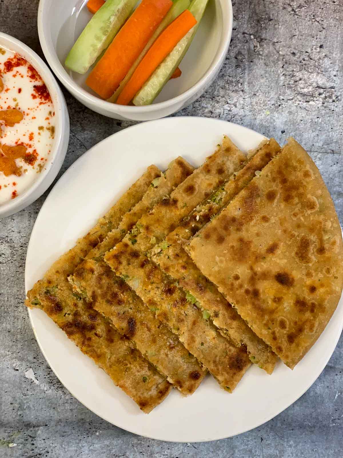 Stuffed Broccoli Paneer Paratha served on a plate with raita and cut cucumber and carrots on the side