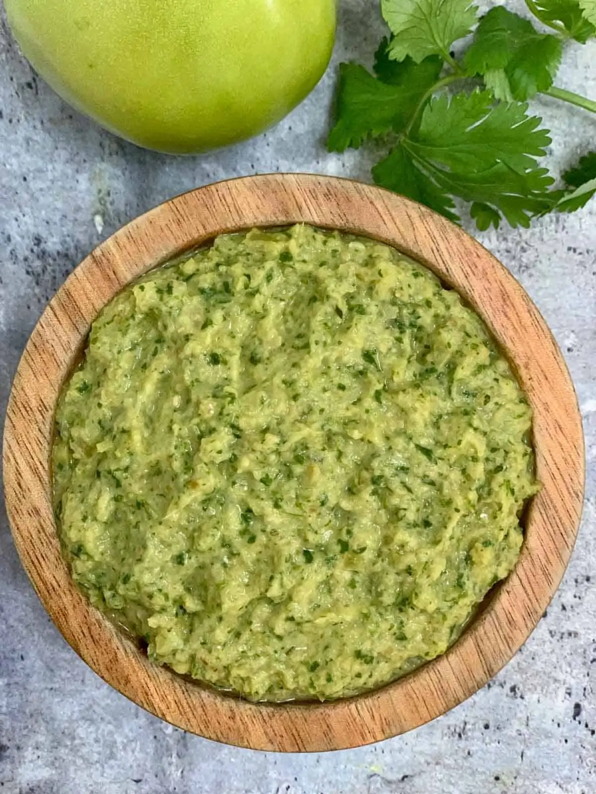 green tomato chutney served in a wooden bowl with cilantro and raw tomato on the side