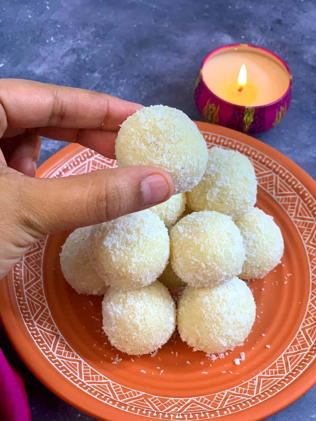 Holding a coconut ladoo made with condensed milk in a finger with the other laddus placed on the plate and candle is lit on the side.
