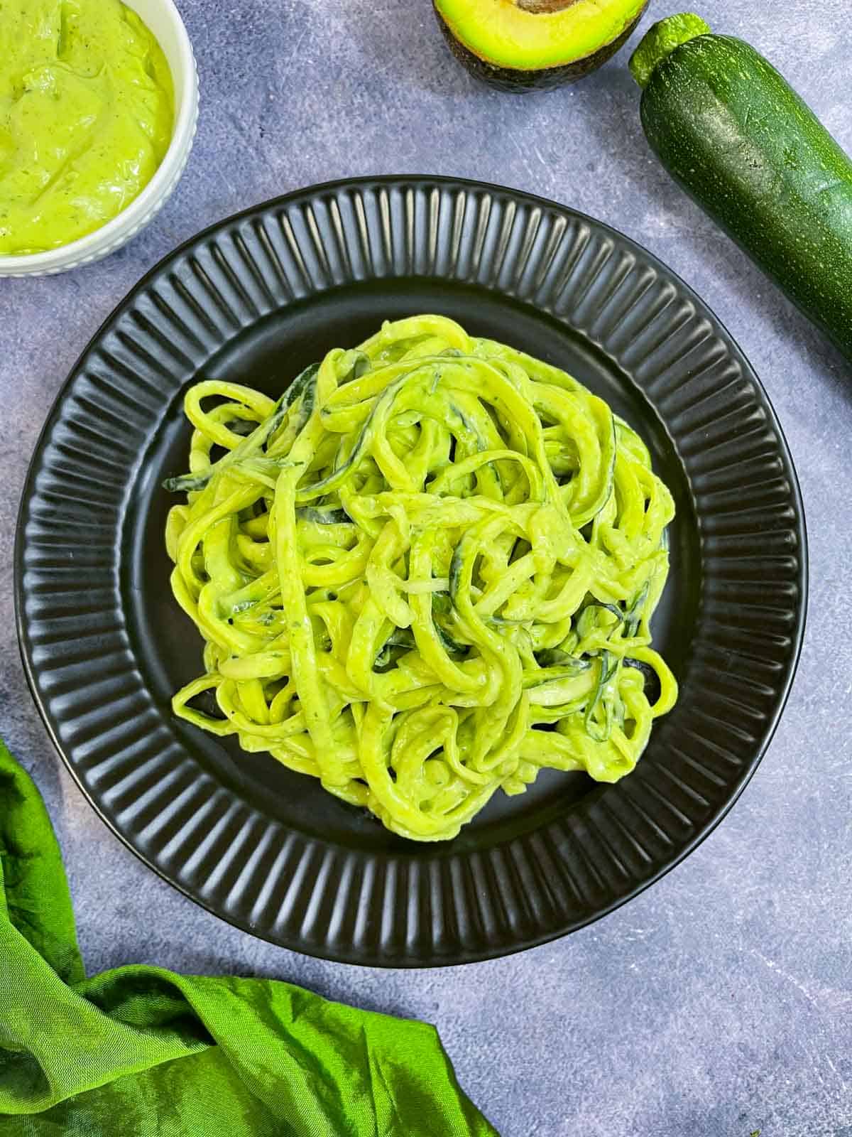 Zucchini Pasta with avocado sauce served in a black plate with cut avocado, zucchini and sauce in the side