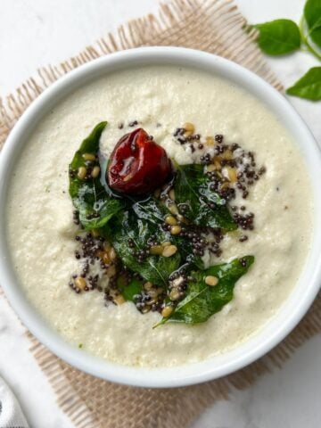 coconut chutney served in a white bowl with tempering on the top and curry leaves on the side