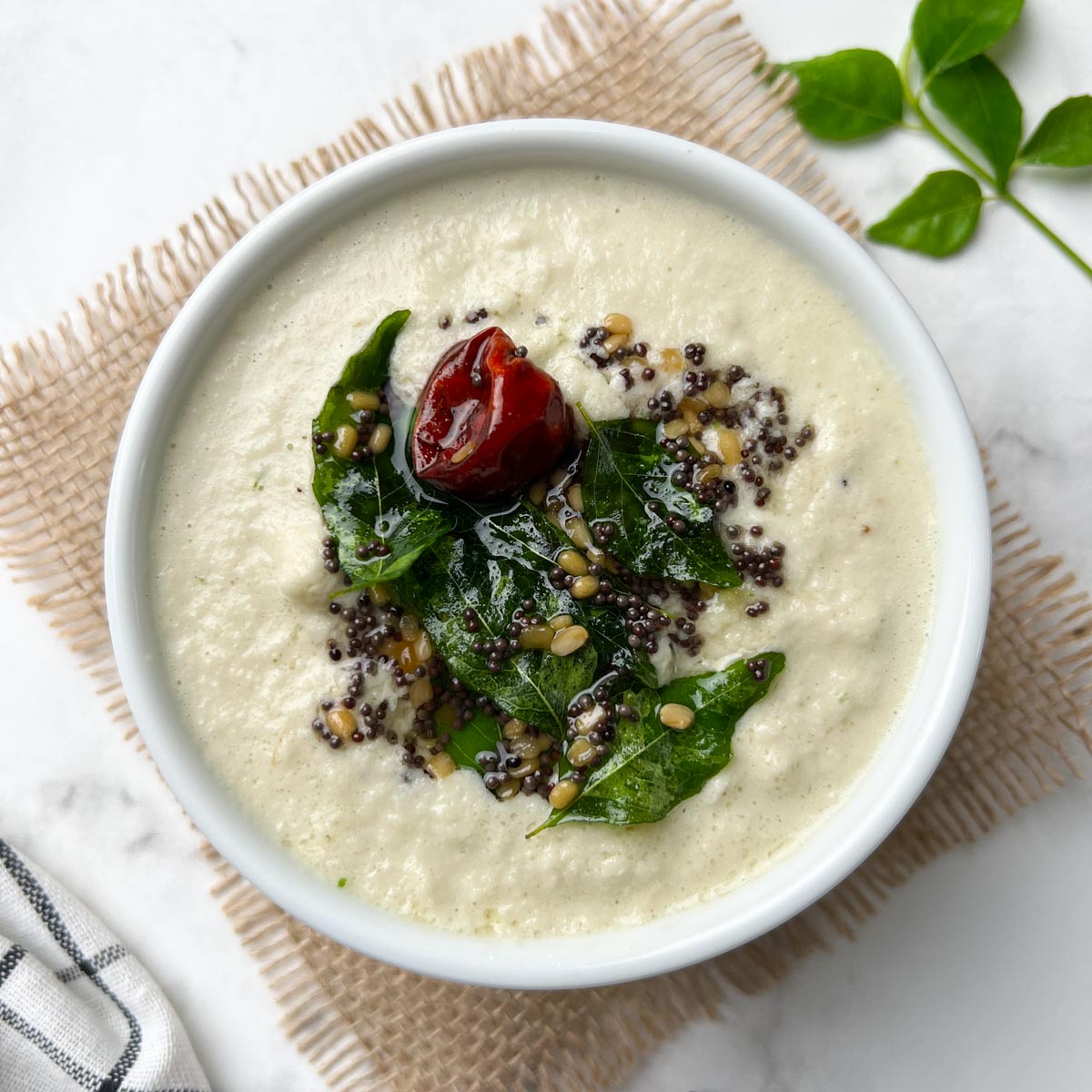 coconut chutney served in a white bowl with tempering on the top and curry leaves on the side