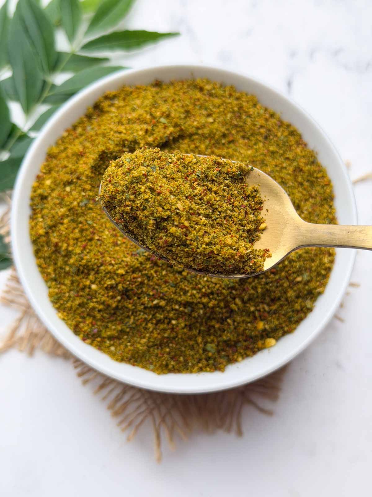 Curry leaves chutney powder in a white bowl with a spoon full of podi and curry leaves stalks on the side for decoration.