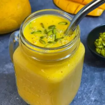 fresh mango milkshake served in a mason jar garnished with pistachios on top with a steel straw and mangoes on the side
