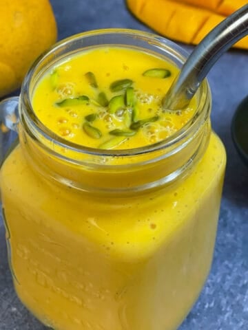 fresh mango milkshake served in a mason jar garnished with pistachios on top with a steel straw and mangoes on the side