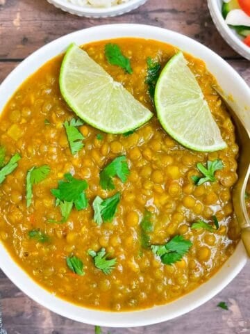 whole masoor dal served in a bowl garnished with lemon wedges and cilantro with a spoon and white rice and salad on the side