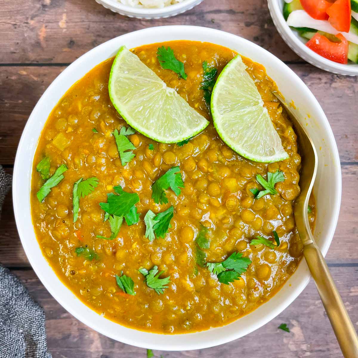 whole masoor dal served in a bowl garnished with lemon wedges and cilantro with a spoon and white rice and salad on the side