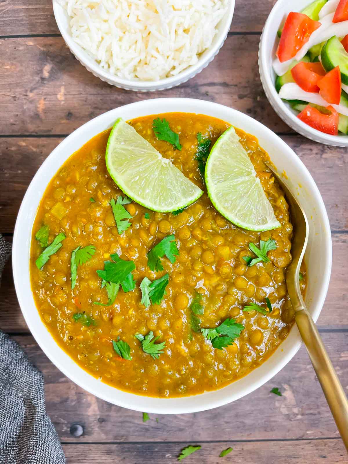 whole masoor dal served in a bowl garnished with lemon wedges and cilantro with a spoon and white rice and salad on the side