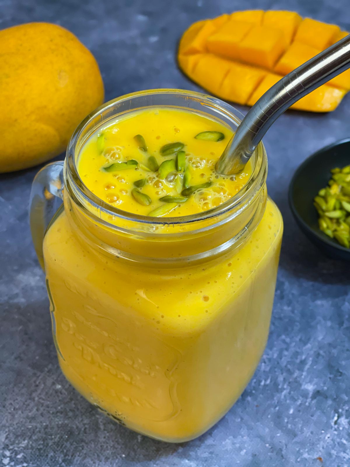 fresh mango milkshake served in a mason jar garnished with pistachios on top with a steel straw and mangoes on the side