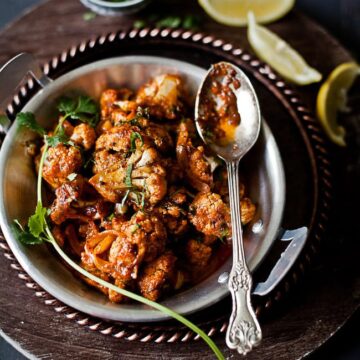 Achari gobi served in a kadai with a spoon and lemon wedges and coriander on the side.