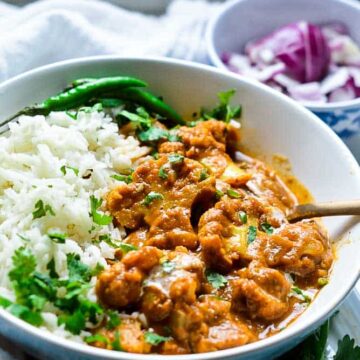 Cauliflower tikka masala served with rice in a bowl with a spoon and raw onion on the side.