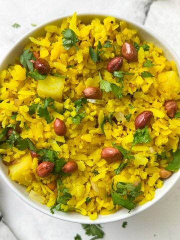 poha recipe served in a white bowl garnished with coriander leaves and roasted peanuts spoon on the side