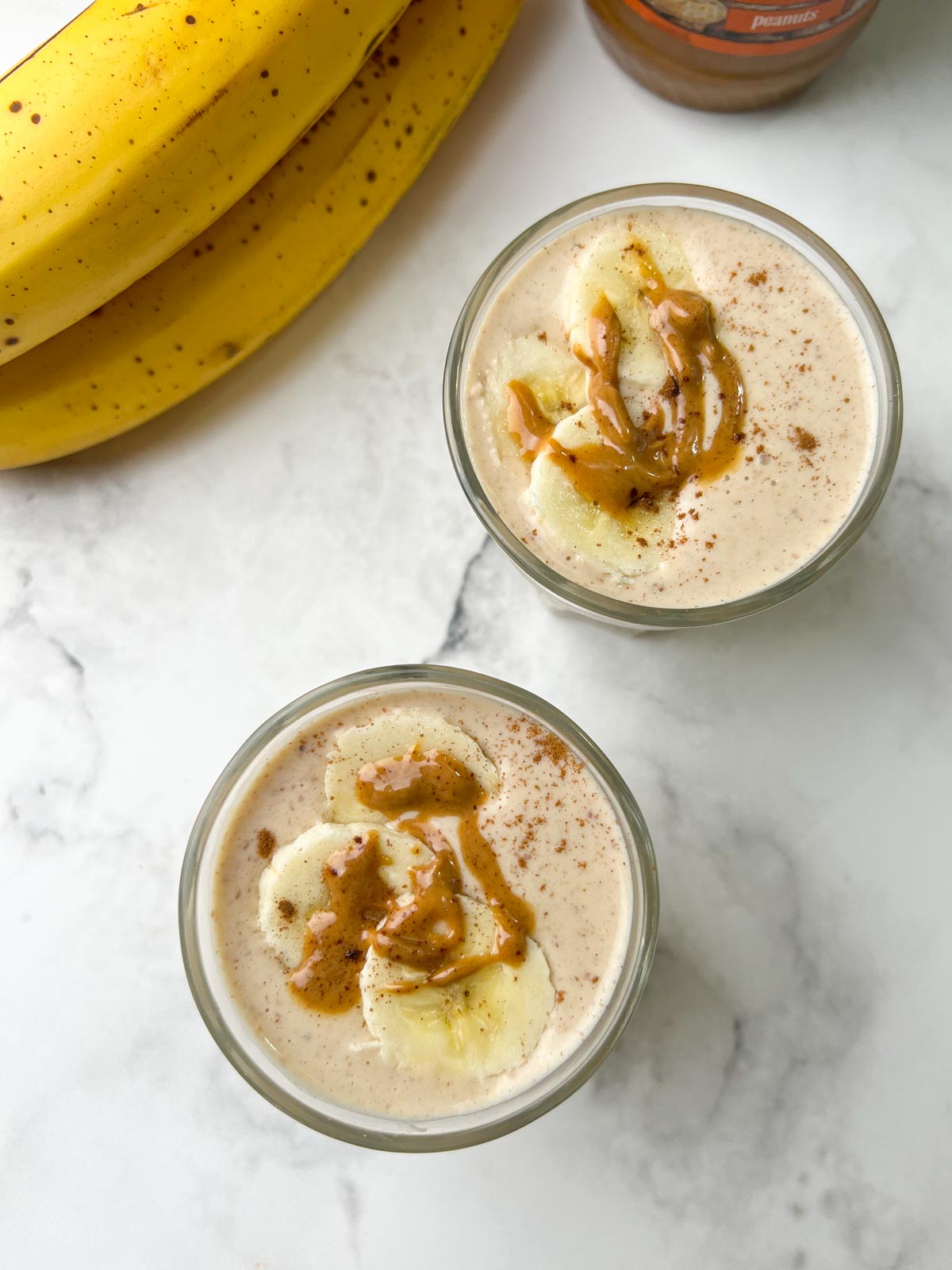 banana peanut butter smoothie served in two glasses toped with banana slices, nut butter and banana on the side
