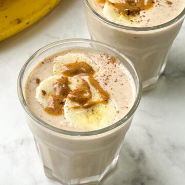 banana peanut butter smoothie served in two glasses toped with banana slices, nut butter and banana on the side
