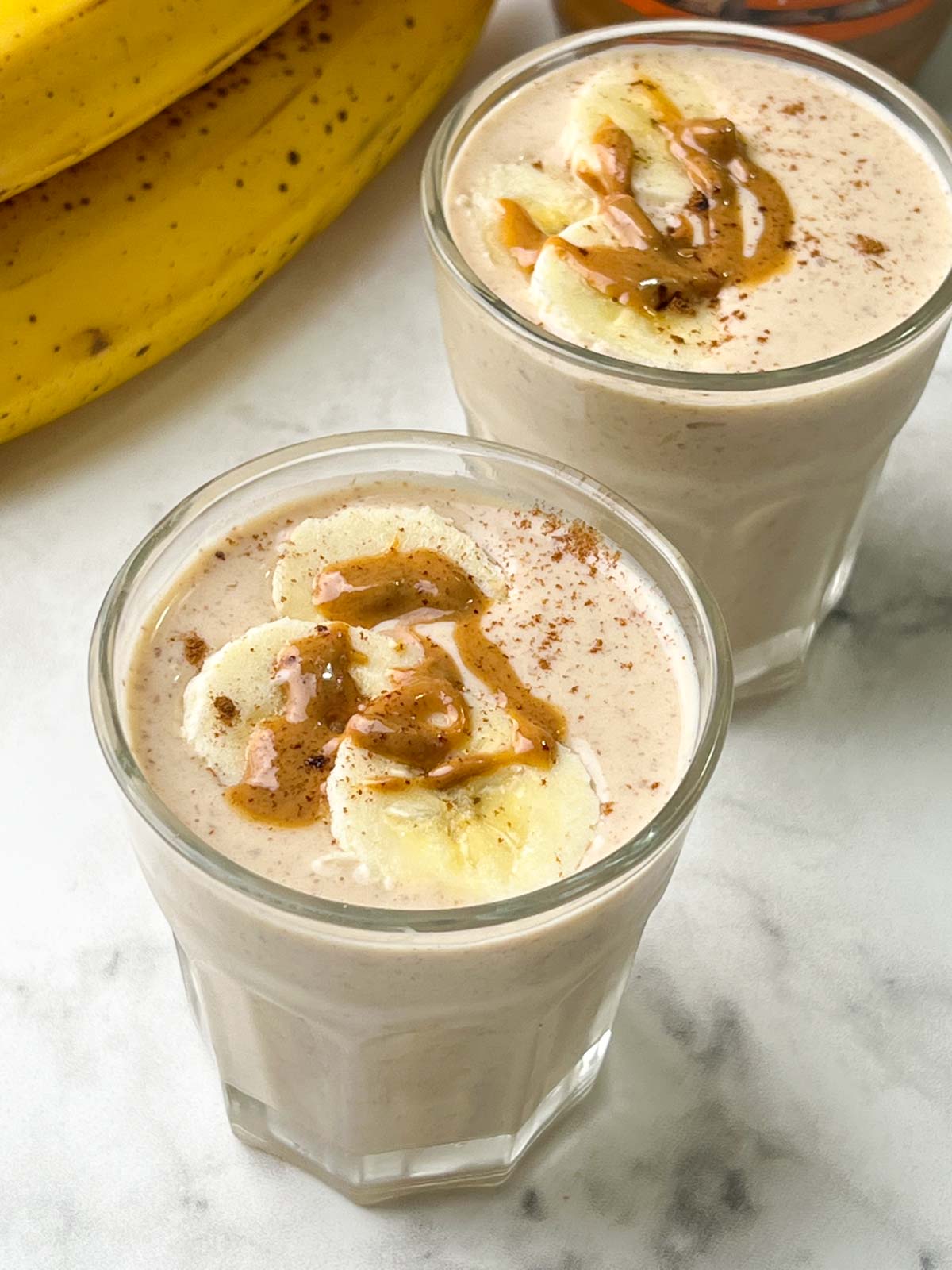 banana peanut butter smoothie served in two glasses toped with banana slices, nut butter and banana on the side