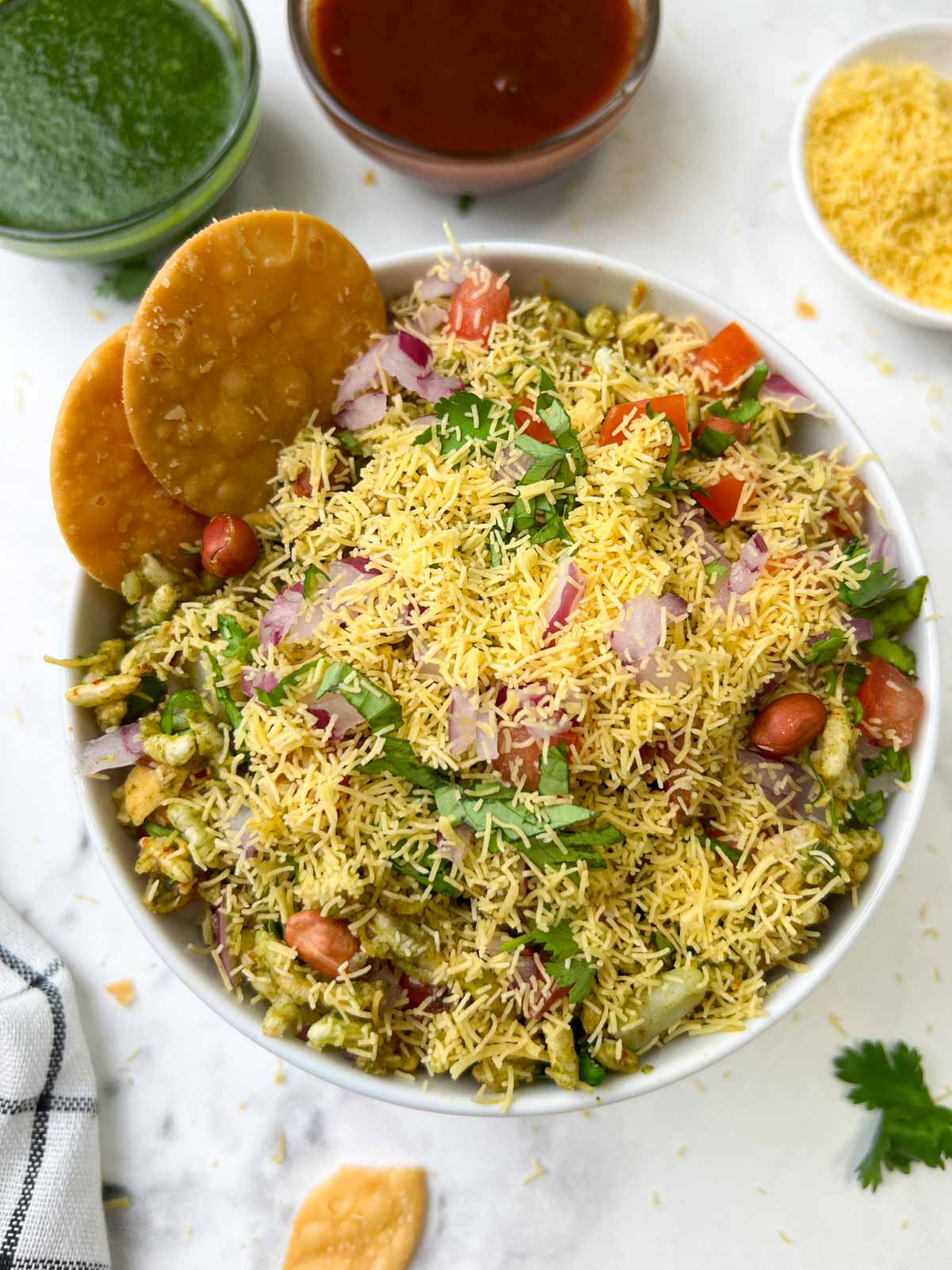 bhel puri chaat served in a white bowl garnished with sev and papdi on the side with green chutney and tamarind chutney on the side