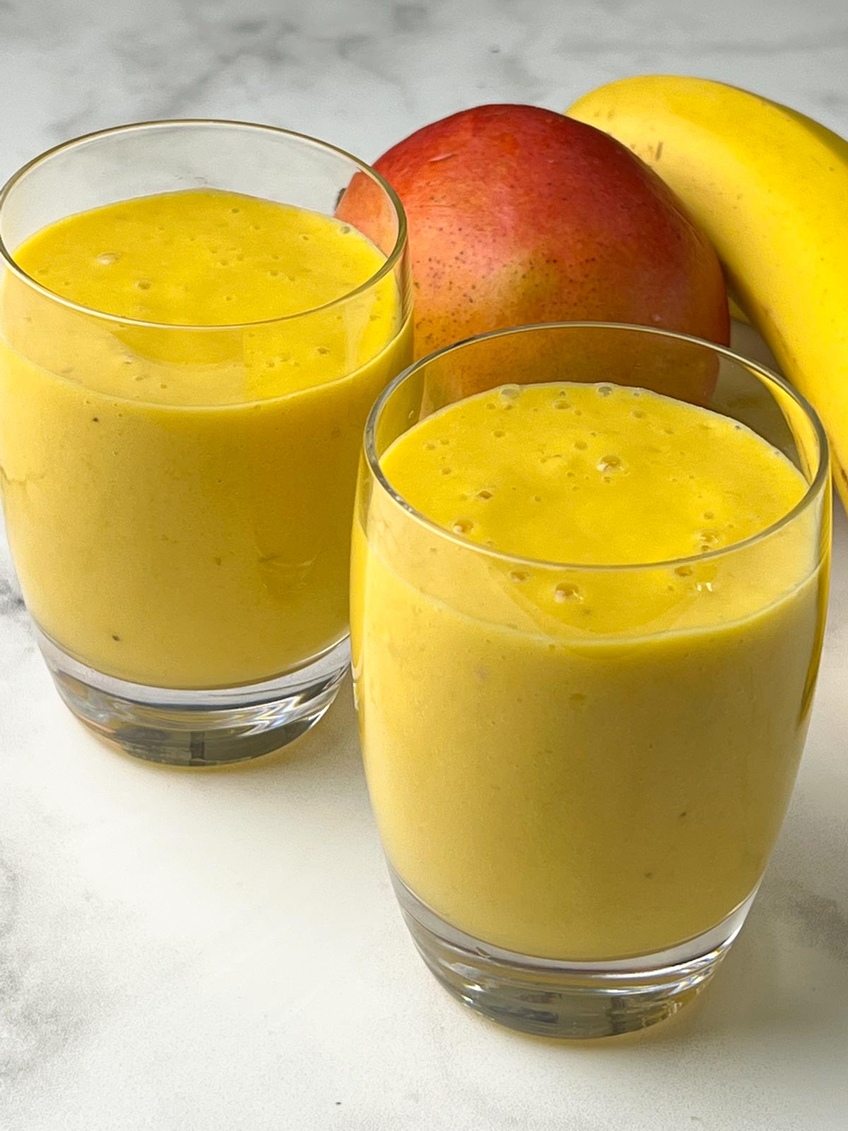 mango banana smoothie served in a serving glasses with whole mango and banana on the side