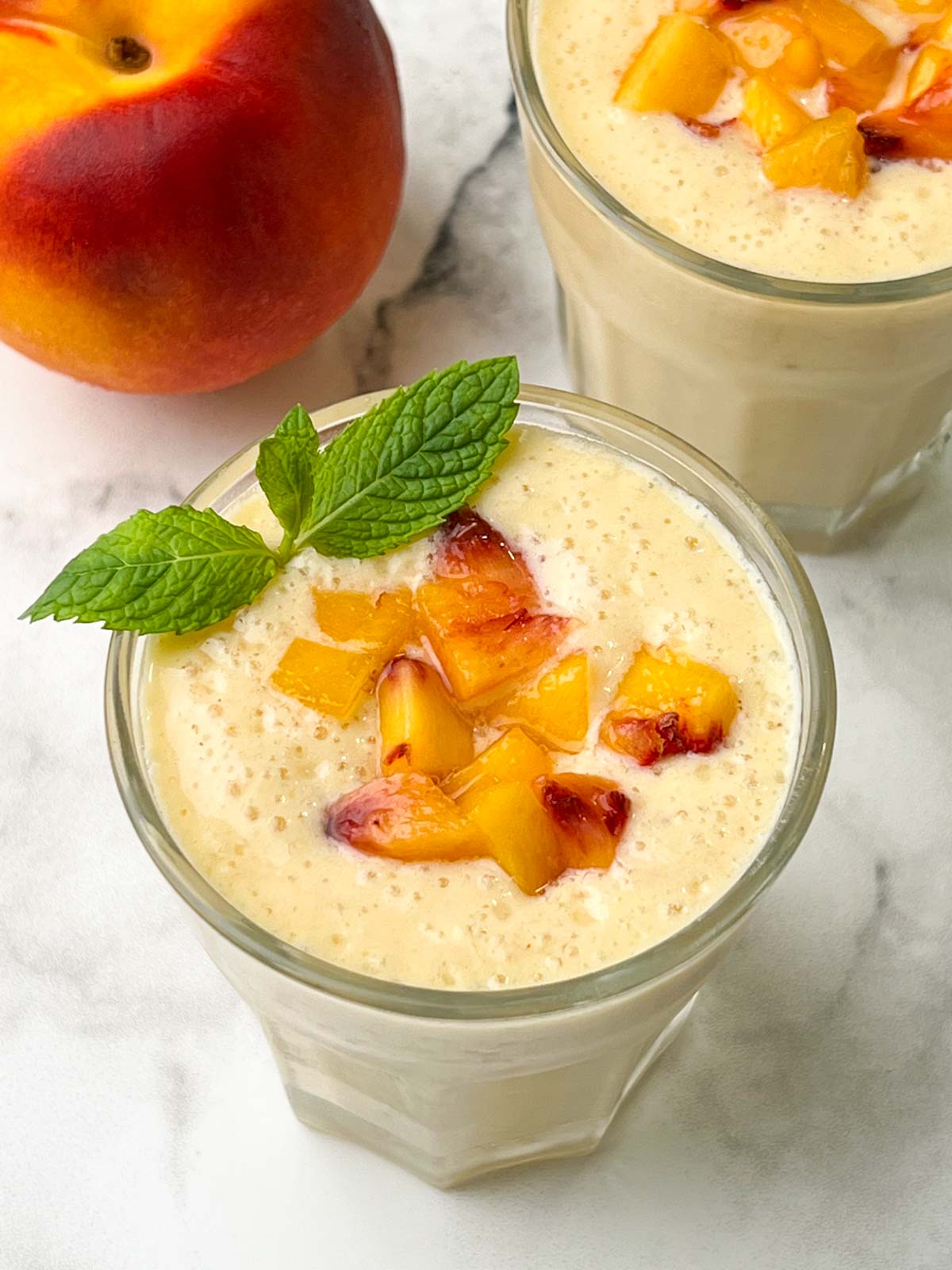 peach banana smoothie served in juice serving glasses topped with chopped yellow peach pieces and garnished with mint leaf
