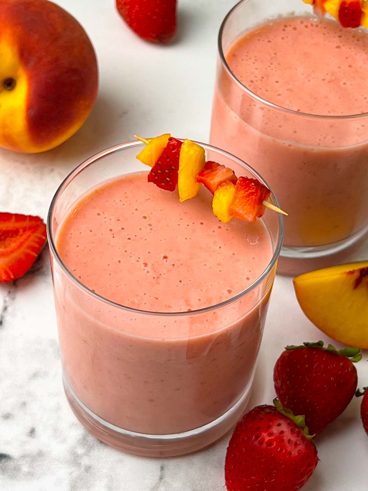 strawberry peach smoothie in juice glasses garnished with peach and strawberry skewers with fruits on the side