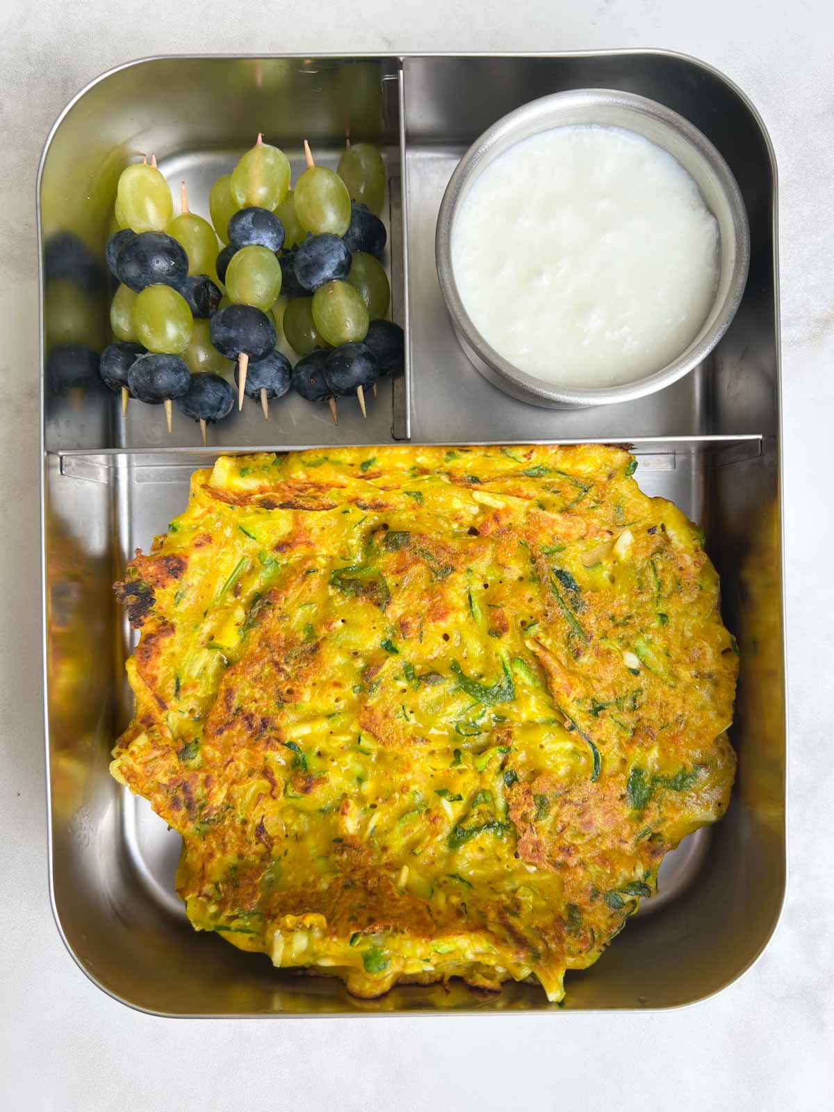 Zucchini Paneer Cheela/Pancake, Yogurt, Fruits in steel bento lunch box.