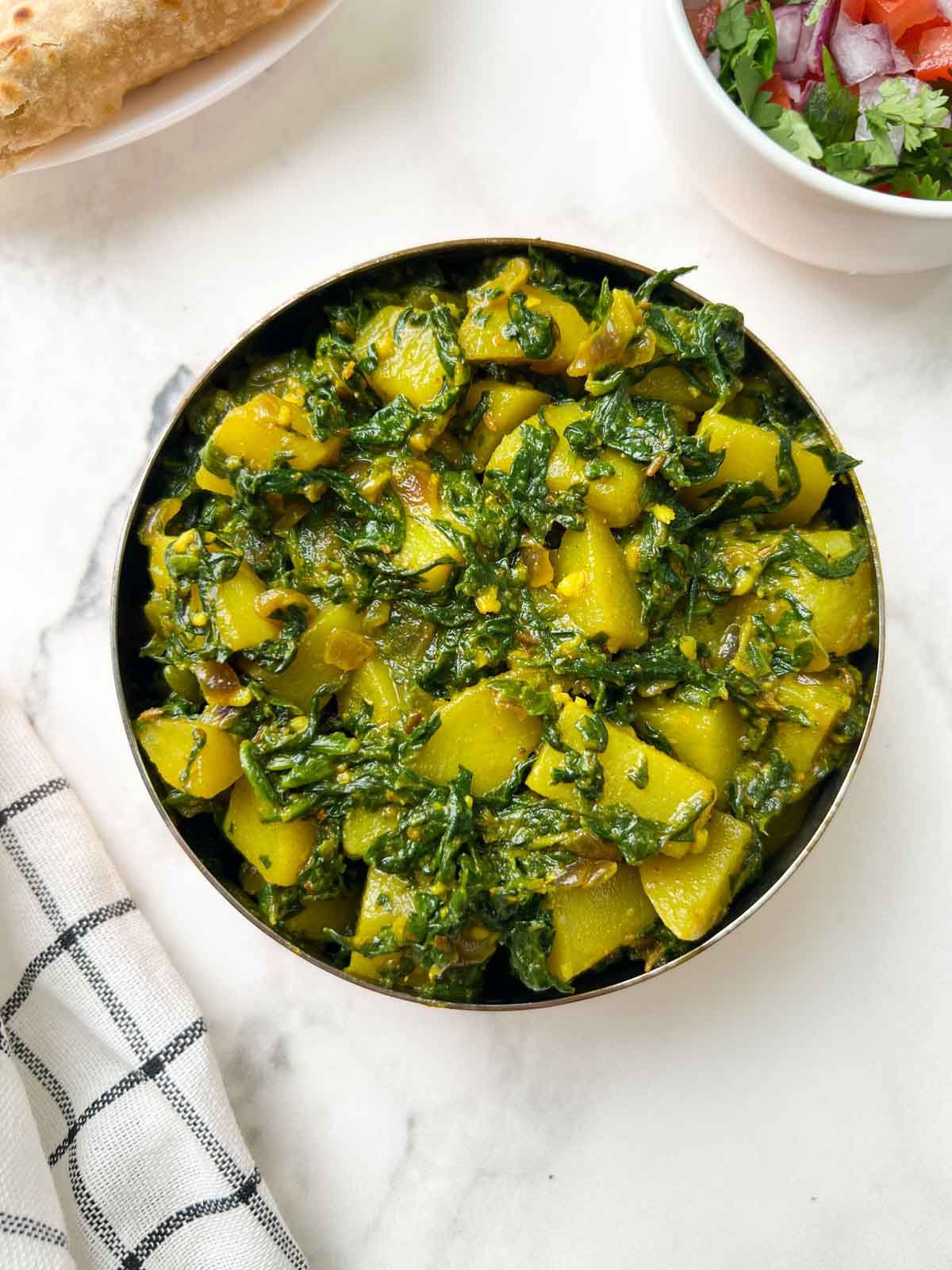 potato spinach stir fry served in a bowl with chapati and salad on the side