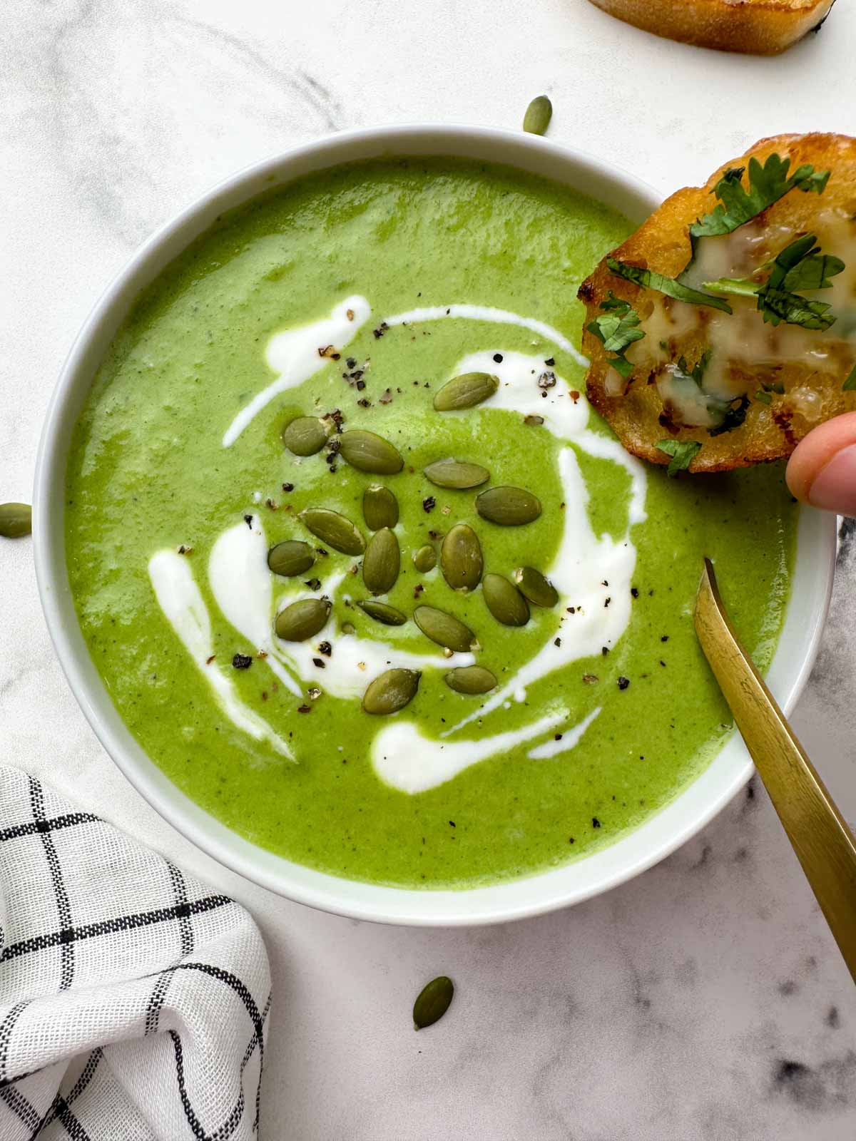 broccoli spinach soup served in a bowl with spoon garnished with cream and pumpkin seeds with garlic bread on the soup