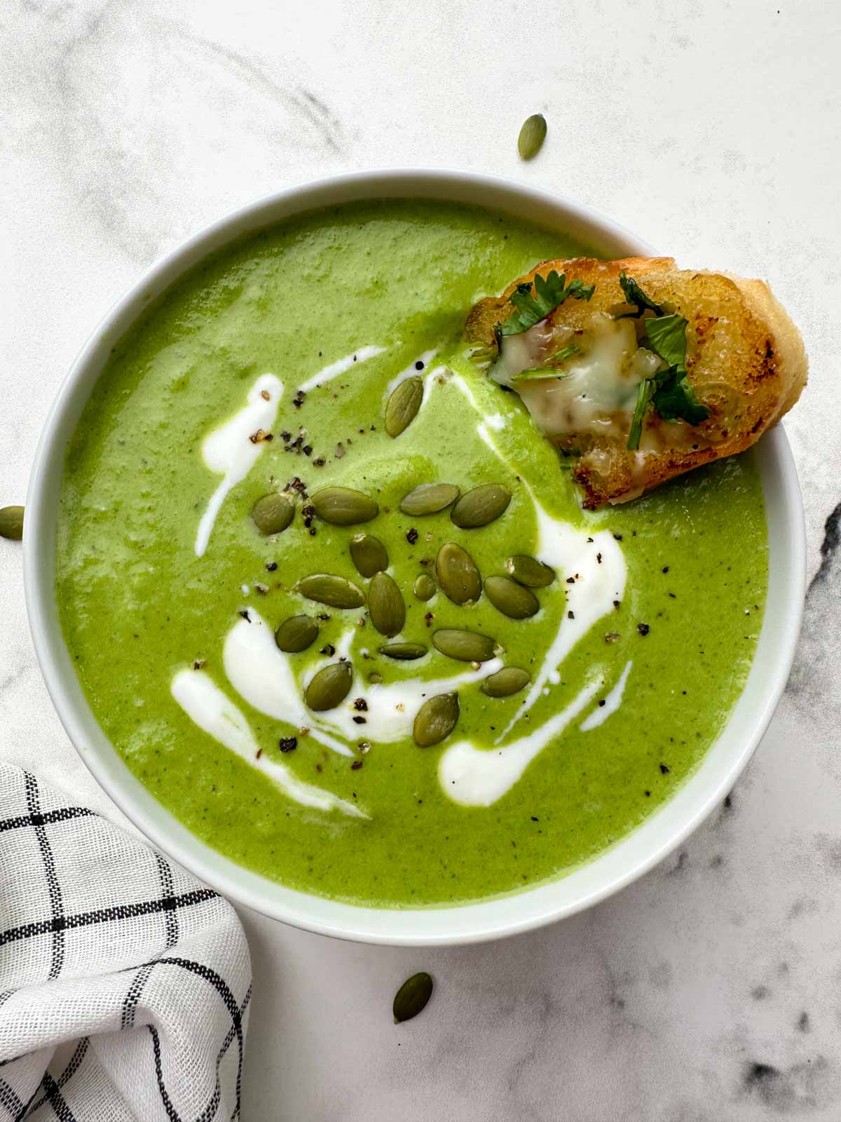 broccoli spinach soup served in a bowl garnished with cream and pumpkin seeds with garlic bread on the soup