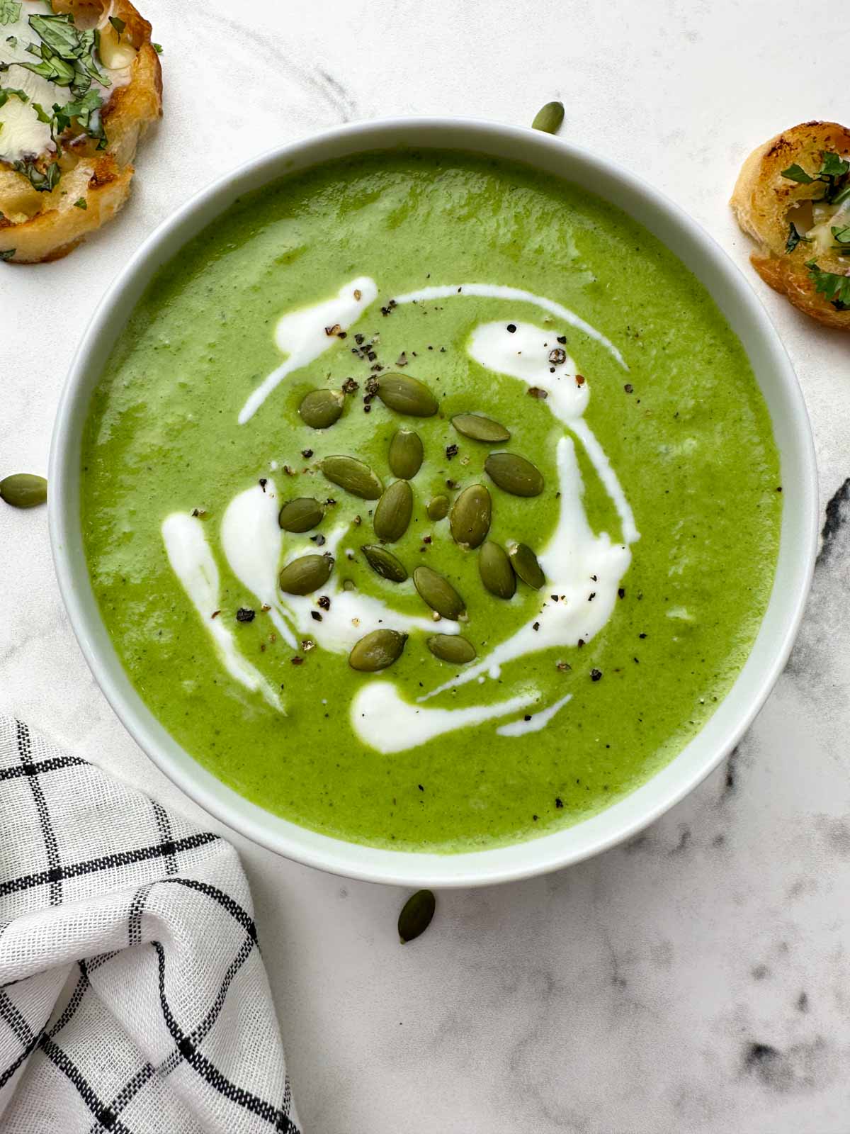 broccoli spinach cheese soup served in a bowl garnished with cream and pumpkin seeds with garlic bread on the side