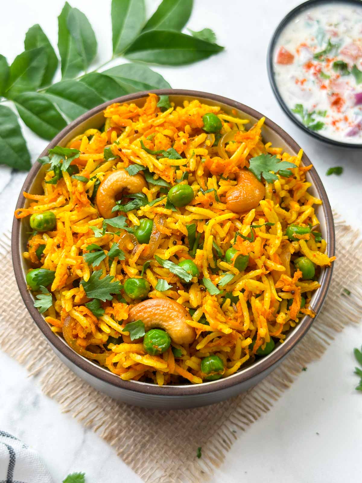 Indian carrot pulao served in a bowl with raita on the side along with spices and curry leaves