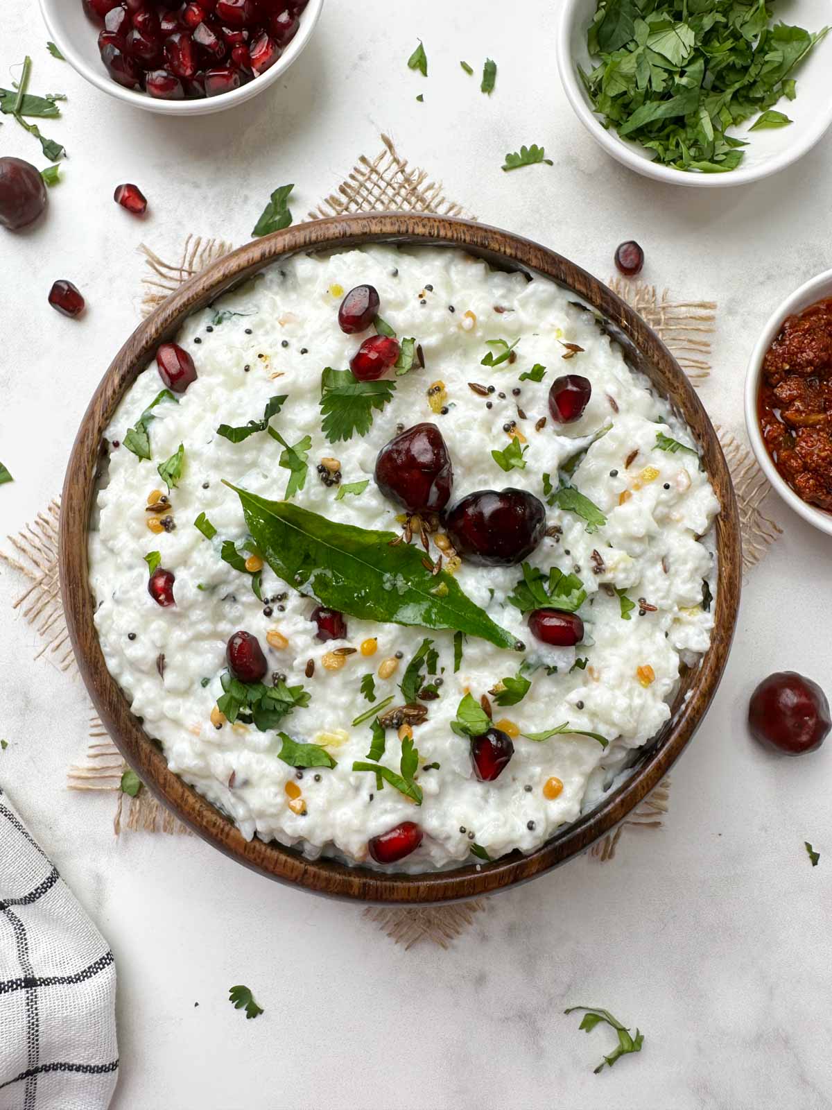 curd rice (thayir saddam) served in a wooden bowl garnished with the tempering with pomegranate seeds, coriander leaves and pickle on the side
