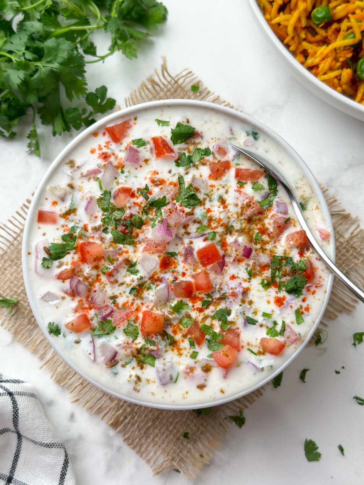 onion tomato raita recipe served in a bowl garnished with roasted cumin powder, red chili powder with a spoon
