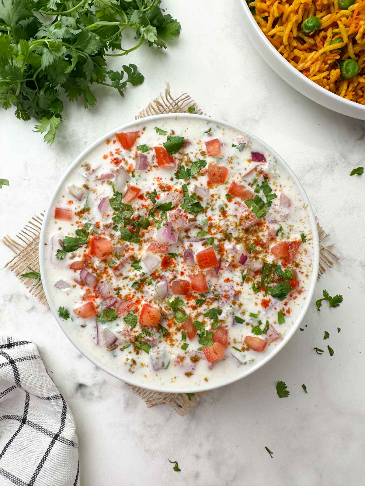 onion tomato raita served on a bowl garnished with roasted cumin powder, red chili powder with carrot rice and coriander bunch on the side