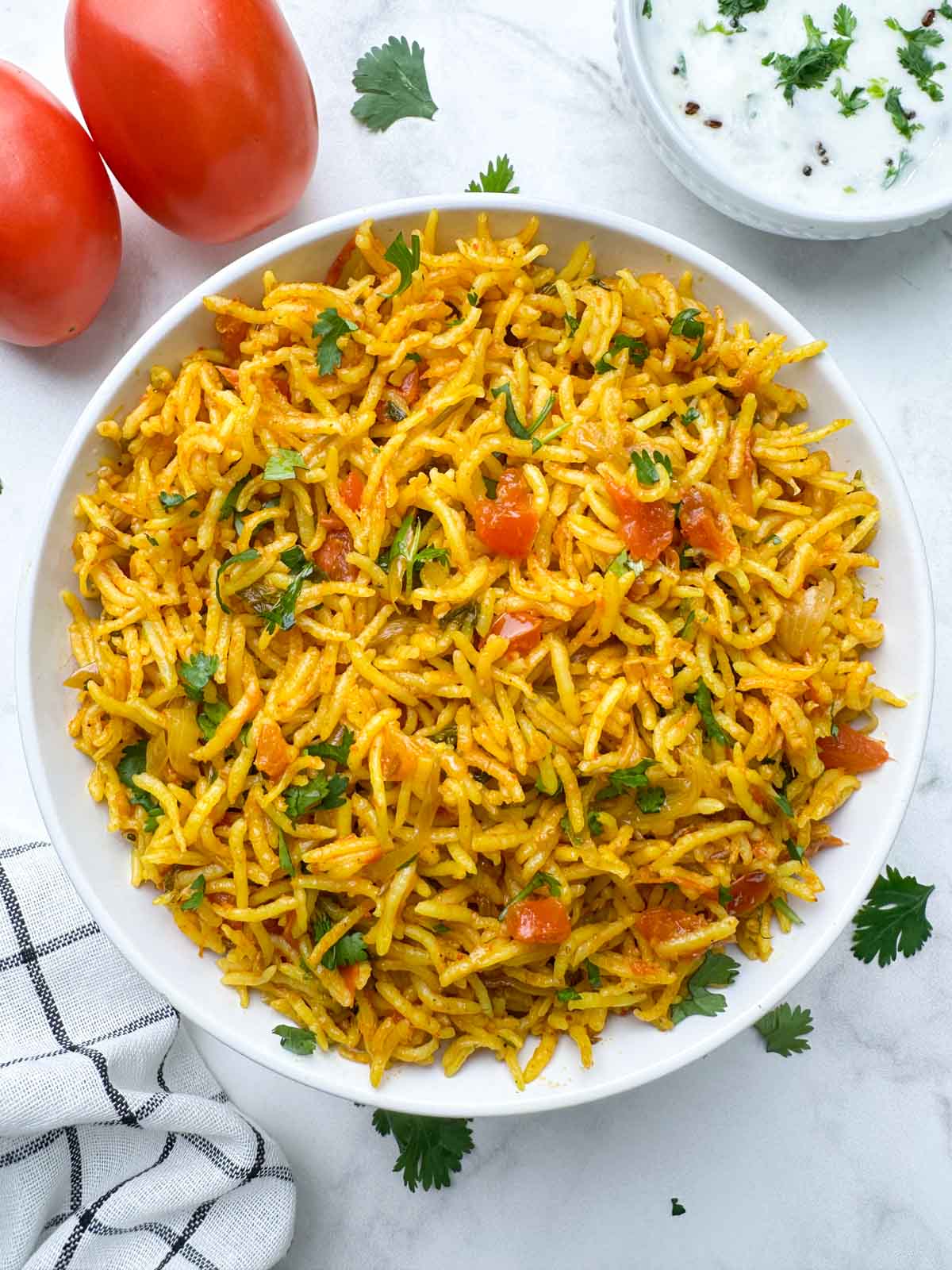 tomato rice made in coconut milk served in a bowl with raita and 2 whole tomatoes on the side