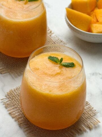 cantaloupe (muskmelon) juice served in 2 serving glasses with mint on the top and a bowl of cut cantaloupe pieces