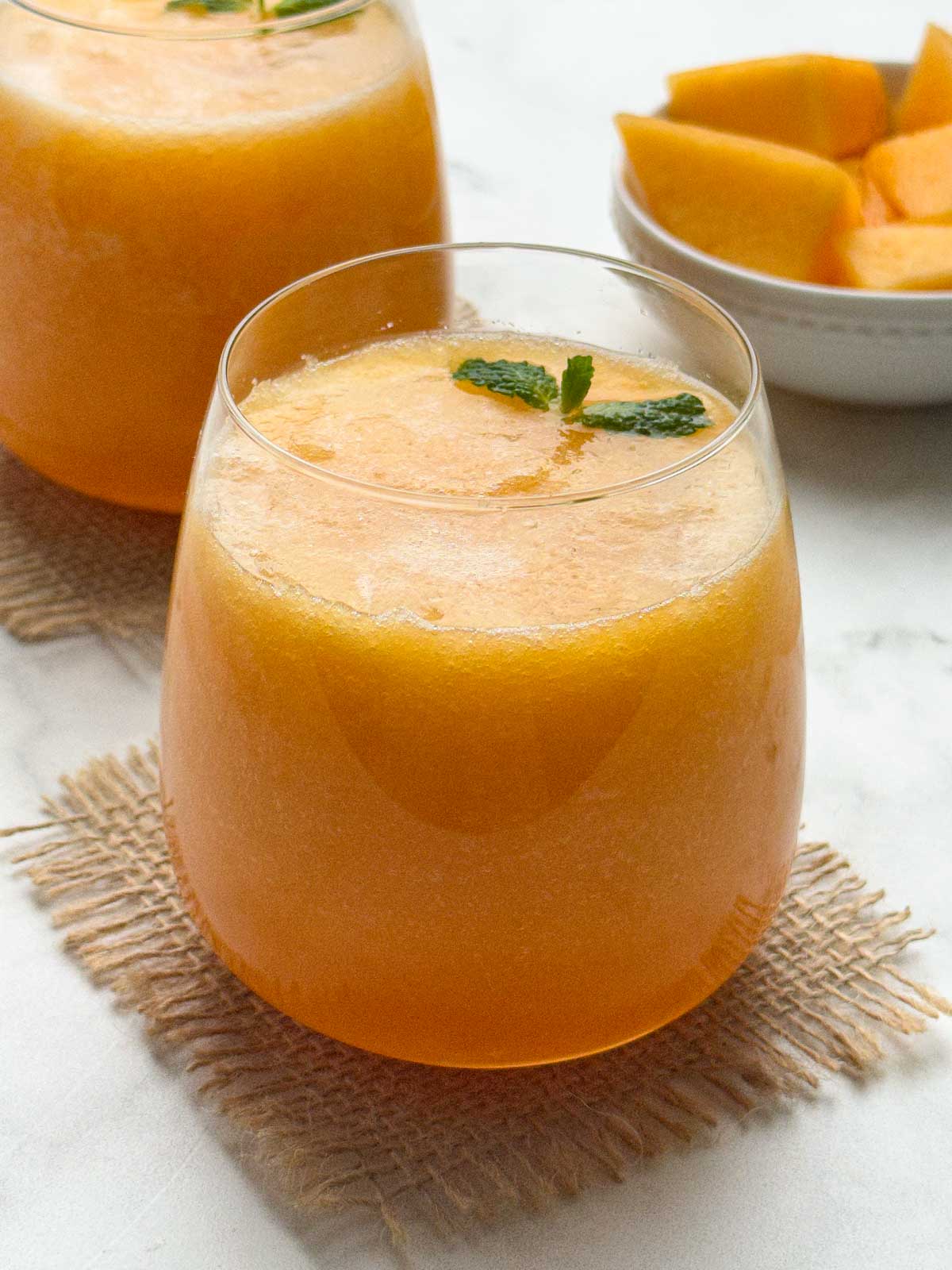 cantaloupe (muskmelon) juice served in 2 serving glasses with mint on the top and a bowl of cut cantaloupe pieces