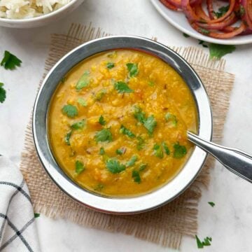 restaurant style dal fry served in a bowl garnished with coriander leaves with a steel spoon and rice and onion salad on the side
