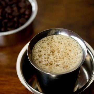 South indian filter coffee served in a steel bowl with coffee beans on the side.