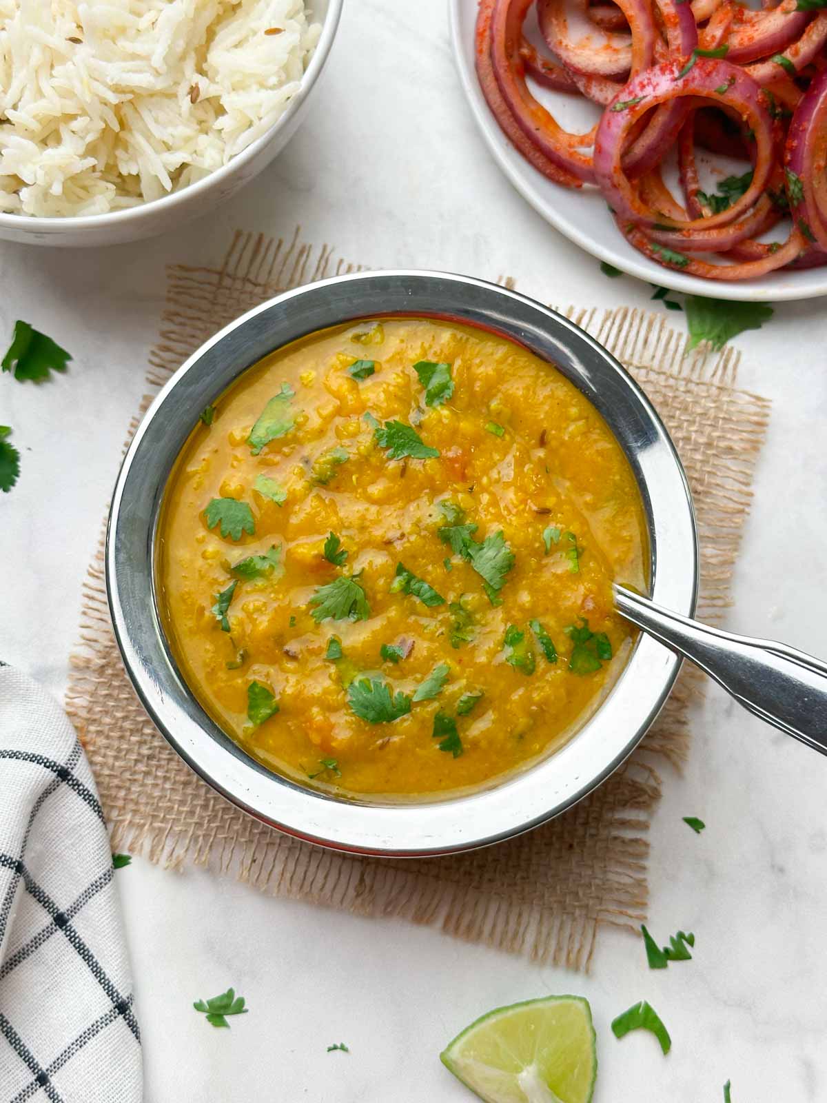 restaurant style dal fry recipe served in a bowl garnished with coriander leaves with a steel spoon and rice and onion salad on the side