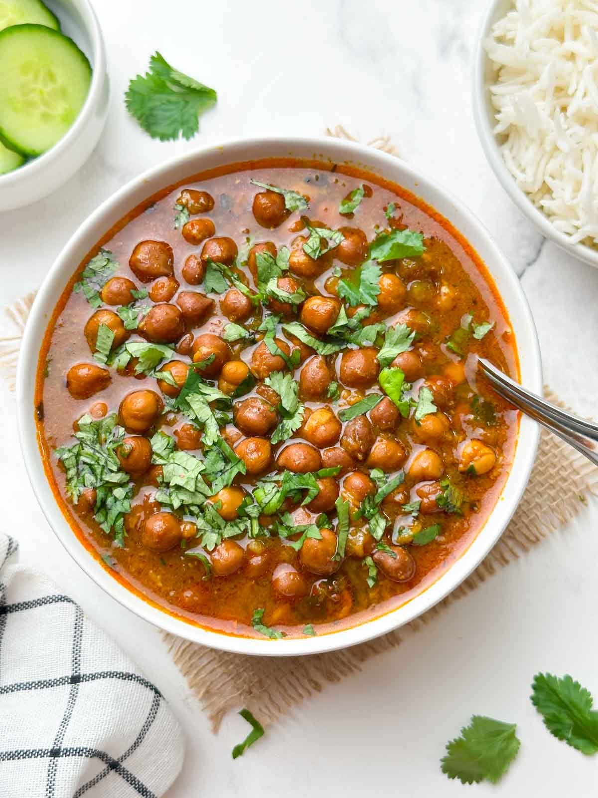 black chickpeas curry (punjabi kala chana) served in a bowl garnish with coriander leaves and spoon in the bowl with cucumber and jeera rice on the side