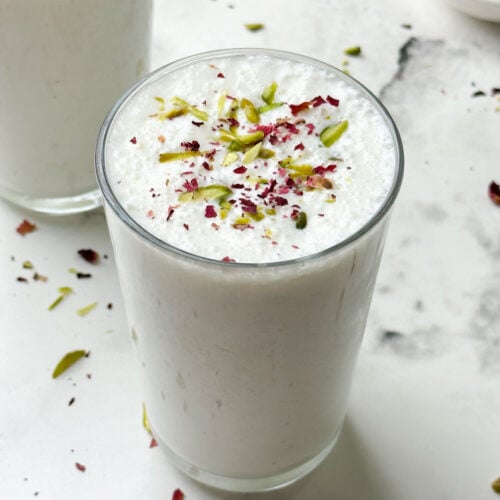 two glass of sweet lassi served in serving glasses garnished with pistachios and dried rose petals