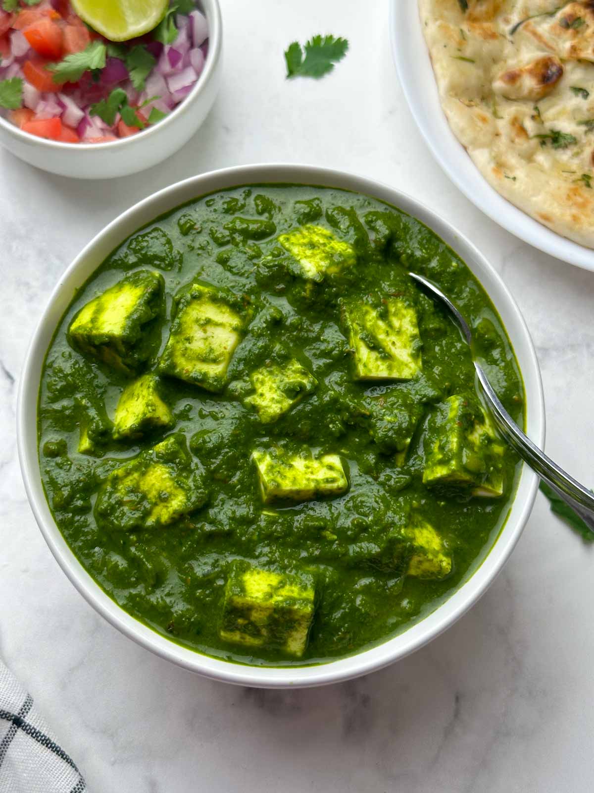 palak paneer served in a white bowl with a spoon and salad and garlic naan on the side