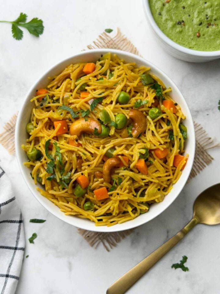 vermicelli upma served in a white bowl with a spoon and green coconut chutney on the side