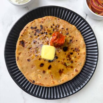 aloo paratha stacked on a plate with butter and pickle on the top with yogurt, laccha onion on the side