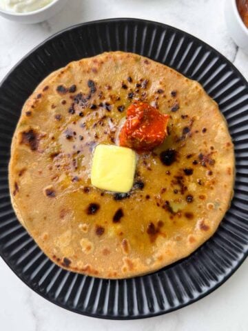aloo paratha stacked on a plate with butter and pickle on the top with yogurt, laccha onion on the side