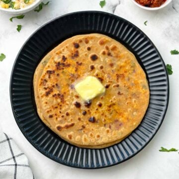 stack of paneer parathas on a black plate with butter on top and raita and pickle on the side