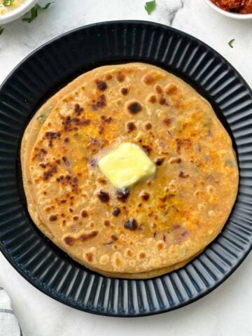 stack of paneer parathas on a black plate with butter on top and raita and pickle on the side