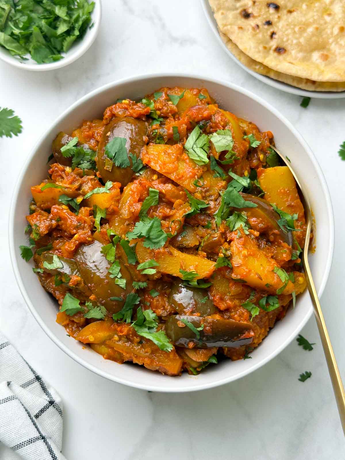 aloo baigan served in a bowl with a spoon with roti and coriander leaves on the side