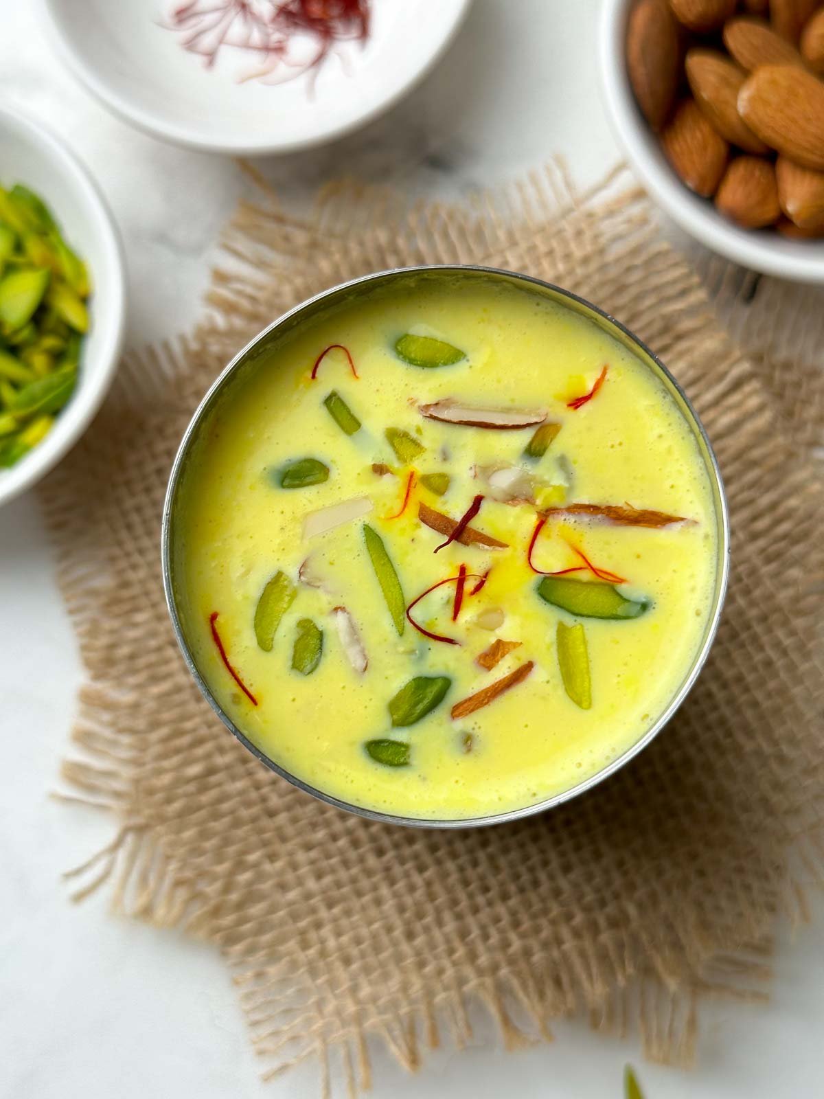 badam milk served in a steel glass garnished with nuts and saffron with nits on the side